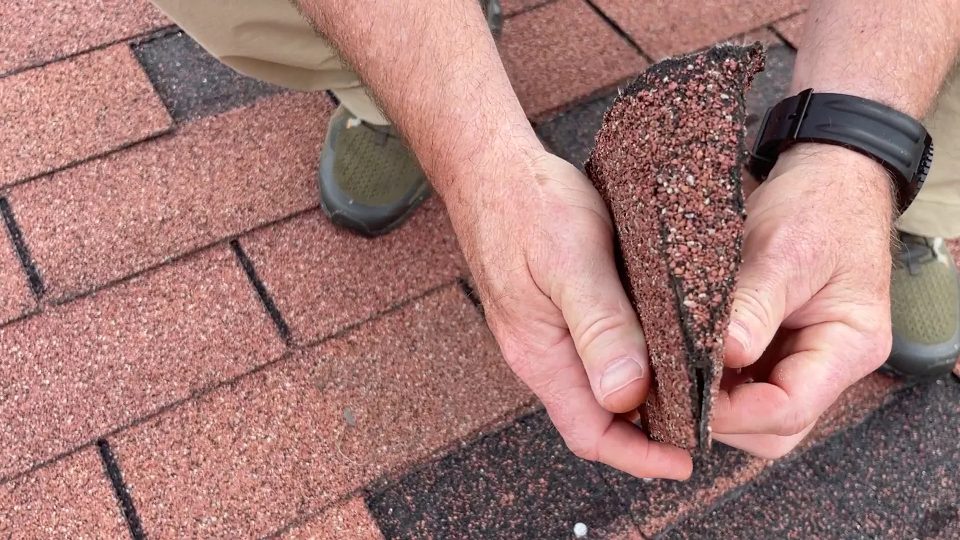 Close-up of hands bending an asphalt shingle to show wear, brittleness, and loss of flexibility on an aging roof.